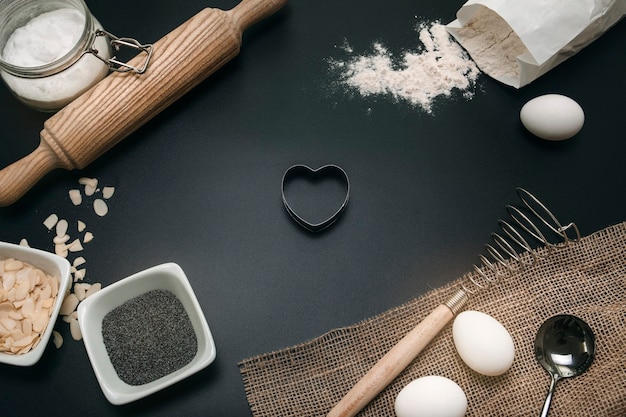 Backen Liebe. Bäckerei Hintergrund. Backzutaten und Küchenutensilien auf schwarzem Hintergrund. Mehl, Mandelnüsse, Eier. Ansicht von oben, flach. Moderne Kochkomposition.