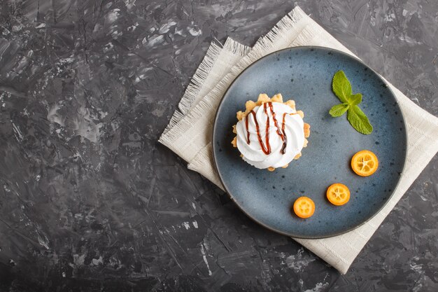 Backen Sie mit Schlagsahne auf einer blauen Keramikplatte mit Scheiben der japanischen Orange und tadellosen Blättern auf einem schwarzen Beton zusammen. Draufsicht, Kopie, Raum.