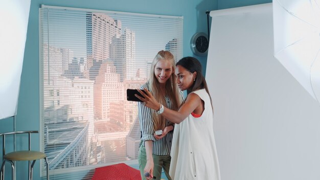 Backstage da sessão de fotos: atraente modelo negra fazendo selfie com linda maquiadora. Eles estão sorrindo e se divertindo.