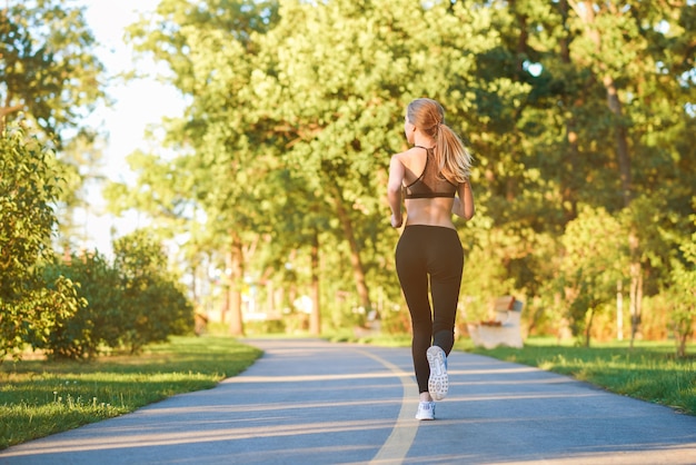 Backview de chica deportiva corriendo a lo largo de la pista de carreras en el parque de la ciudad.