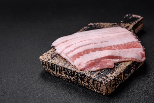Bacon cru fresco cortado em fatias com especiarias salgadas e ervas em um fundo de concreto texturizado escuro