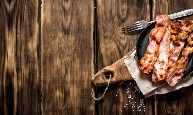 Bacon frito em uma frigideira com um garfo sobre uma mesa de madeira