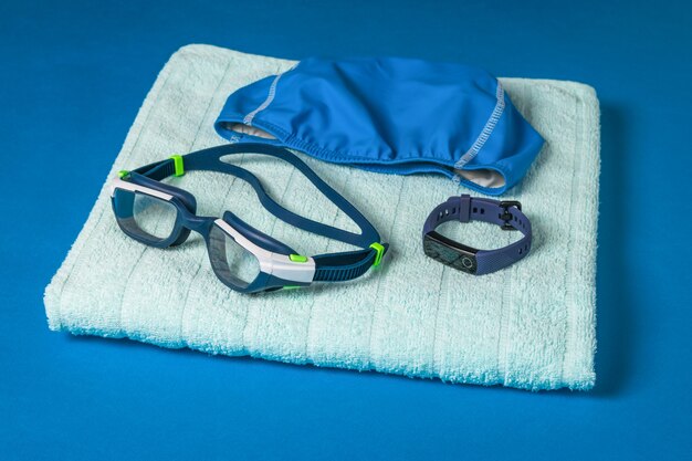 Badekappe, Brille und schickes Armband auf einem Handtuch auf einer blauen Oberfläche. Zubehör zum Schwimmen im Pool.