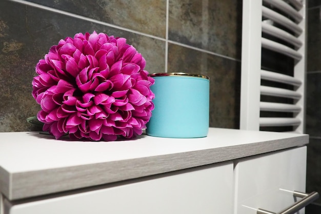 Badezimmer-Dekorelemente Ein blaues Cremeglas und eine rosa leuchtende künstliche Doppelblume auf einem Schrankregal Schwarze Wandfliesen Badezimmer- und Toiletteninnenraum Heizkörper-Handtuchwärmer an der Wand
