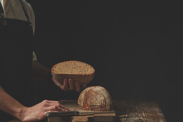 Bäcker, der auf dunklem Hintergrund eine Hälfte frisch gebackenes Brot in der Hand hält