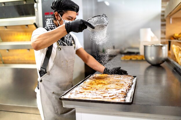 Bäcker streut Zucker auf einen Kuchen in einer Bäckerei und trägt eine Gesichtsmaske