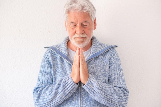 Bärtiger älterer Mann in Freizeitkleidung mit gefalteten Händen, die vor weißem Hintergrund betend stehen Älterer Mann im blauen Pullover im geistlichen Gebet