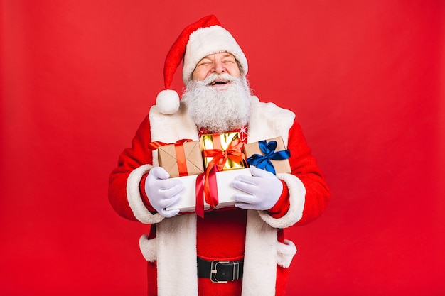 bärtiger alter Mann im Weihnachtsmannkostüm, das Geschenke hält, die lokal auf rotem Hintergrund stehen