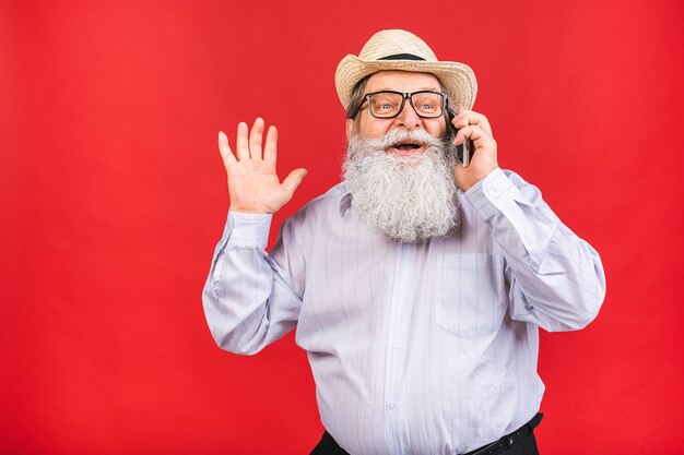 bärtiger alter Mann mit Hut und Brille, die auf einem Handy lokalisiert auf rotem Hintergrund sprechen