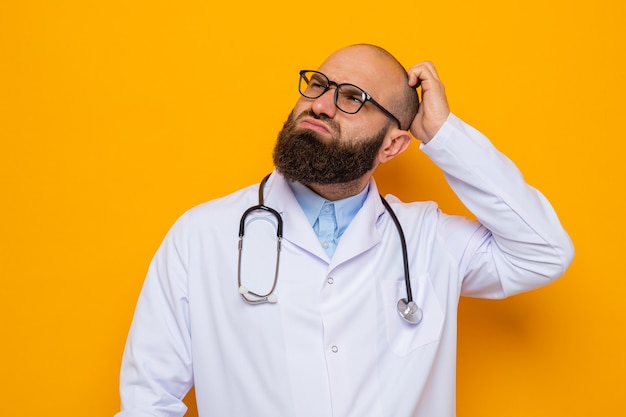 Bärtiger Arzt im weißen Kittel mit Stethoskop um den Hals mit Brille, der verwirrt aufblickt und sich den Kopf kratzt, der über orangefarbenem Hintergrund steht
