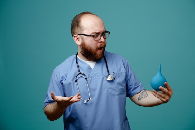 Bärtiger Arzt in Uniform mit Stethoskop um den Hals, der eine Brille mit Einlauf hält und ihn verwirrt und enttäuscht über blauem Hintergrund ansieht