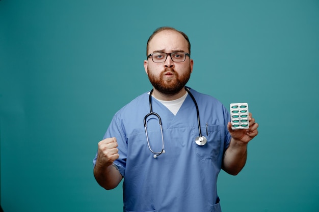 Bärtiger Arzt in Uniform mit Stethoskop um den Hals, der eine Brille mit Pillen trägt und mit ernstem Gesicht und geballter Faust auf blauem Hintergrund in die Kamera schaut