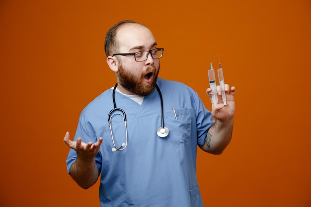 Bärtiger Arzt in Uniform mit Stethoskop um den Hals, der eine Brille mit Spritzen trägt und erstaunt und überrascht über orangefarbenem Hintergrund steht