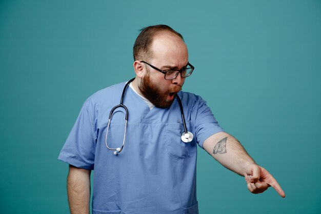 Bärtiger Arzt in Uniform mit Stethoskop um den Hals, der eine Brille trägt und beiseite schaut, überrascht und schockiert ist und mit dem Zeigefinger auf etwas zeigt, das über blauem Hintergrund steht