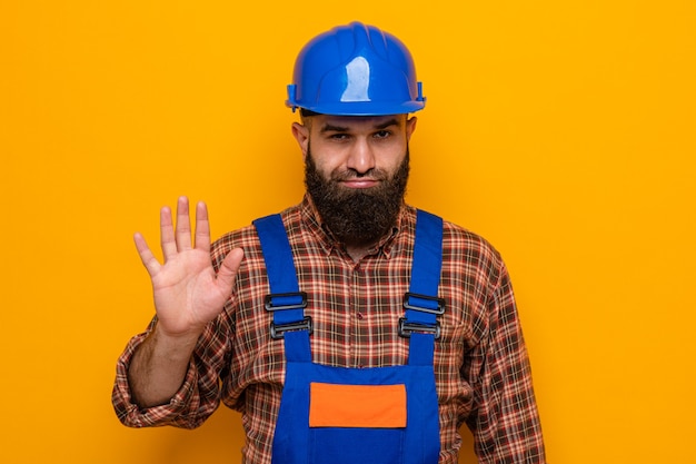 Bärtiger Baumeister in Bauuniform und Schutzhelm, der unzufrieden in die Kamera schaut und eine Stopp-Geste mit der Hand über orangefarbenem Hintergrund zeigt