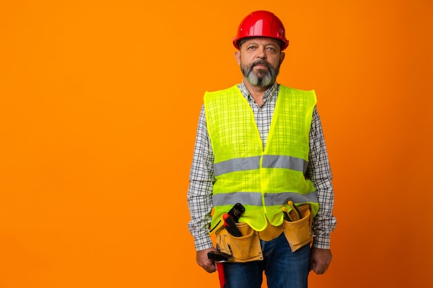 Bärtiger Baumeister mittleren Alters in Uniform und Bauarbeiterhelm mit Werkzeugen vor orangefarbenem Hintergrund