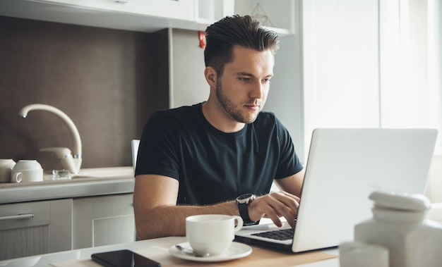 Bärtiger brünetter Mann ist freiberuflich am Laptop zu Hause in der Küche, während er einen Kaffee trinkt