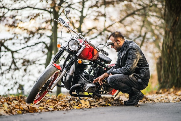 Bärtiger brutaler Mann in Sonnenbrille und Lederjacke, der auf einem Motorrad auf der Straße im Wald sitzt