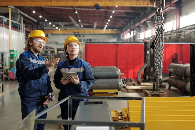 Bärtiger Fabrikingenieur in Bauarbeiterhelm und Schutzbrille, der auf Ketten zeigt und Kollegen erklärt, wie man einen Industriekran benutzt