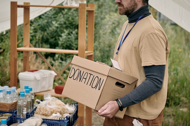 Bärtiger Freiwilliger mit Abzeichen tragender Pappkarton mit Spende zum Tisch mit Waren für Obdachlose