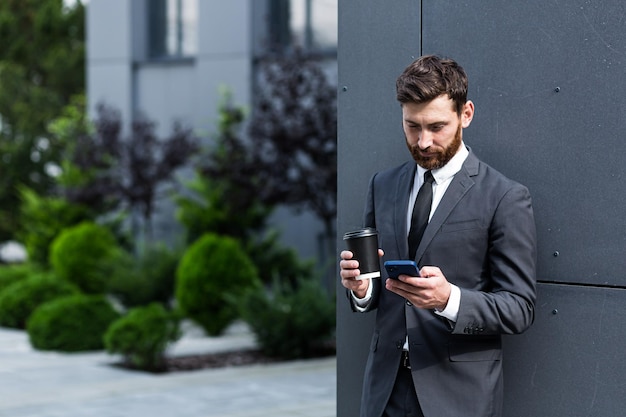 Bärtiger Geschäftsmann im Anzug in der Pause mit Handy verwenden Smartphone. Geschäftsmann, der draußen auf modernem städtischen Straßenhintergrund mit Kaffeetasse in der Innenstadt im Freien steht. Platz kopieren