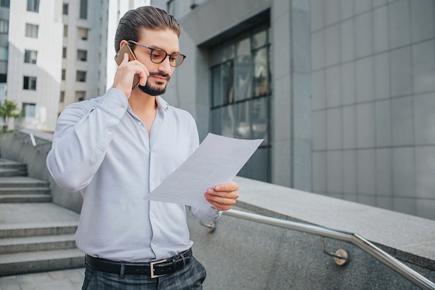 Bärtiger Geschäftsmann mit Sonnenbrille steht auf Stufen und hält ein Stück Papier. Er sieht sich das Dokument an. Guy studiert es. Gleichzeitig telefoniert er.