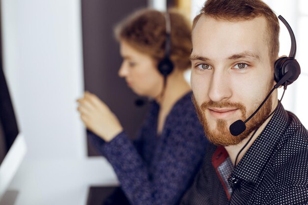 Bärtiger Geschäftsmann spricht mit einem Headset in der Nähe seiner Kollegin, während er in einem modernen, sonnigen Büro sitzt.