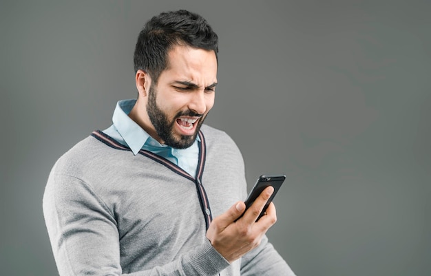 Bärtiger gutaussehender Mann mit wütendem Gesichtsausdruck, der Smartphone in der Hand auf grau isoliertem Hintergrund hält