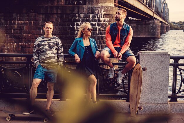 Bärtiger Hepster-Typ mit Longboard und blonde Frau mit Sonnenbrille posiert in der Nähe des Wasserkanals der Stadt.