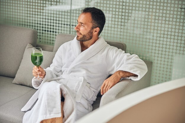 Bärtiger Herr im Bademantel, der auf der Couch sitzt und wegschaut, während er ein Glas frischen Gemüsesaftes hält