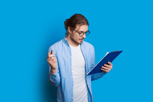 bärtiger Junge mit langen Haaren schaut in ein Notizbuch durch Brille und posiert auf einer blauen Wand