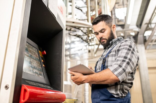 Bärtiger junger Arbeiter der Polymerproduktion, der Daten im Touchpad durchschaut, während er durch das Bedienfeld steht