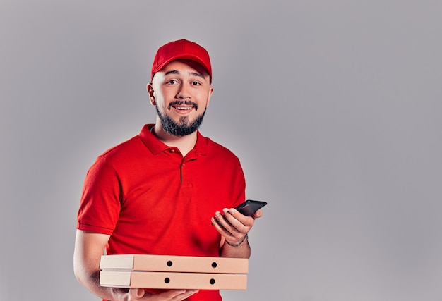 Bärtiger junger Lieferbote in rotem T-Shirt und Mütze mit Pizzakartons und mit Smartphone auf grauem Hintergrund. Schnelle Lieferung nach Hause.