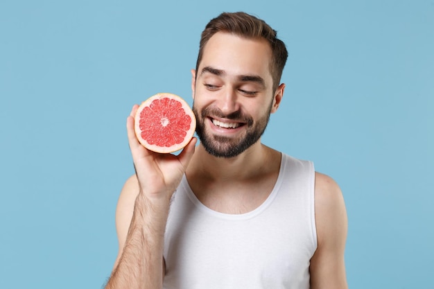 Bärtiger junger Mann 20 Jahre alt in weißem Hemd hält in der Hand die Hälfte der Grapefruit isoliert auf blauem Pastellhintergrund, Studioporträt. Konzept für kosmetische Verfahren im Gesundheitswesen für die Hautpflege. Mock-up-Kopienbereich