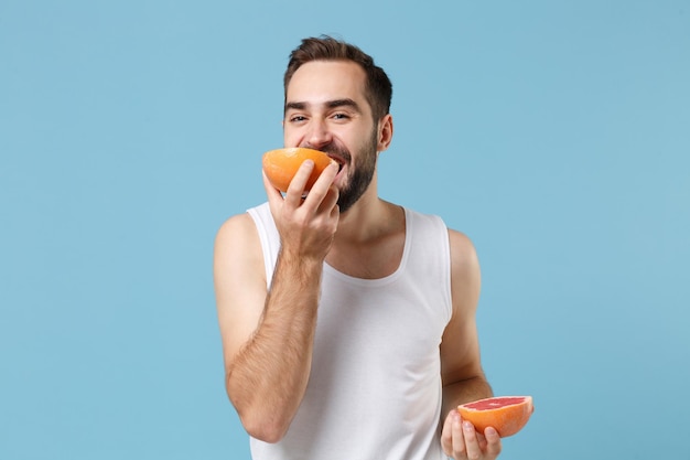 Bärtiger junger Mann 20 Jahre alt in weißem Hemd hält in der Hand die Hälfte der Grapefruit isoliert auf blauem Pastellhintergrund, Studioporträt. Konzept für kosmetische Verfahren im Gesundheitswesen für die Hautpflege. Mock-up-Kopienbereich