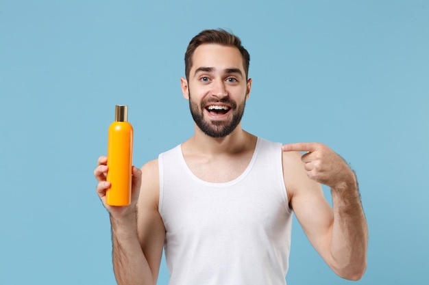 Bärtiger junger Mann im Alter von 20 Jahren in weißem Hemd hält eine Flasche Shampoo isoliert auf blauem pastellfarbenem Wandhintergrund, Studioporträt. Konzept für kosmetische Verfahren im Gesundheitswesen für die Hautpflege. Mock-up-Kopienbereich.