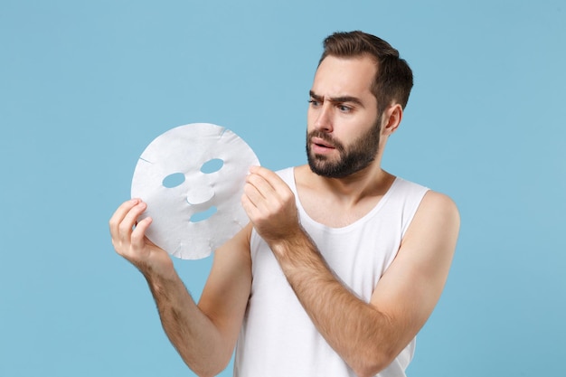 Bärtiger junger Mann im Alter von 20 Jahren in weißem Hemd hält Gesichtsmaske aus Gewebegewebe isoliert auf blauem Pastellhintergrund, Studioporträt. Konzept für kosmetische Verfahren im Gesundheitswesen für die Hautpflege. Mock-up-Kopienbereich.