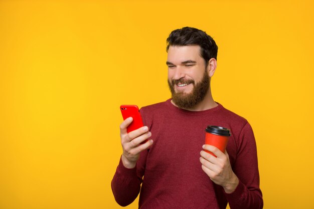 Bärtiger junger Mann im roten Pullover lächelt und hält ein Smartphone und eine Tasse Kaffee auf gelbem Hintergrund.