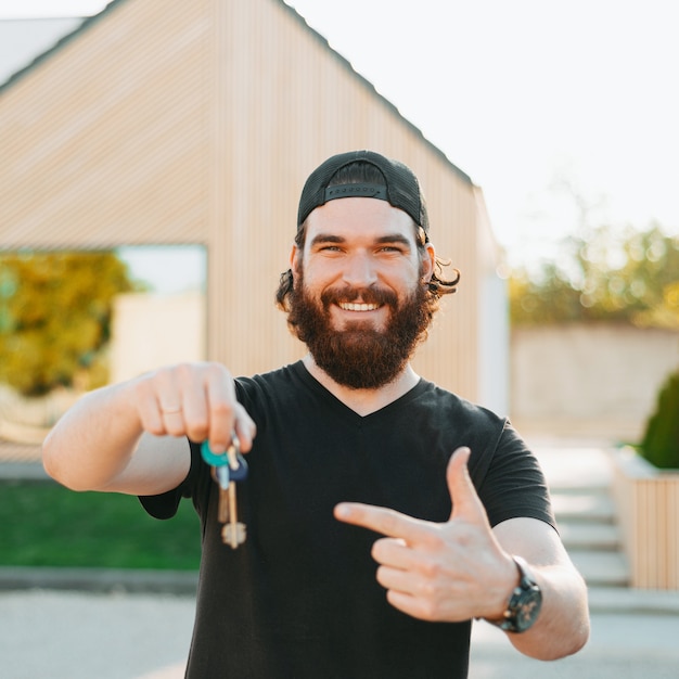 Bärtiger junger Mann lächelt, hält einige Schlüssel und zeigt mit der linken Hand auf sein Haus im Hintergrund