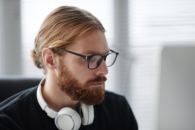 Bärtiger junger Mann mit Brille und Pullover, der auf den Bildschirm des Gadgets blickt