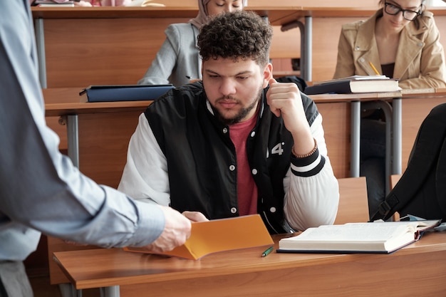 Bärtiger junger Student in Freizeitkleidung, der Notizen in einem Heft durchsieht, das von einem Professor gehalten wird, der ihm erklärt, wie man die Gleichung im Unterricht löst