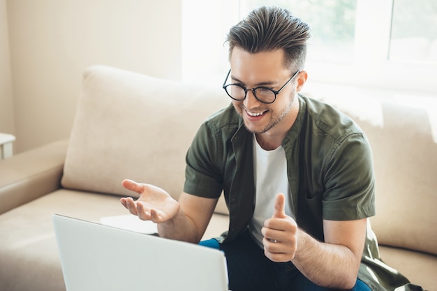 Bärtiger kaukasischer Junge mit Brille, die ein Online-Treffen am Laptop zu Hause sitzt auf Sofa