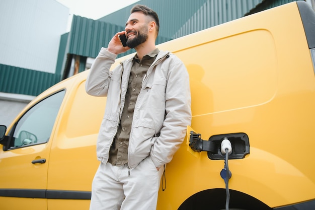 Foto bärtiger kaukasischer mann, der in der nähe eines elektroautos steht, das auflädt und zeitanpassungen auf einem smartphone vornimmt