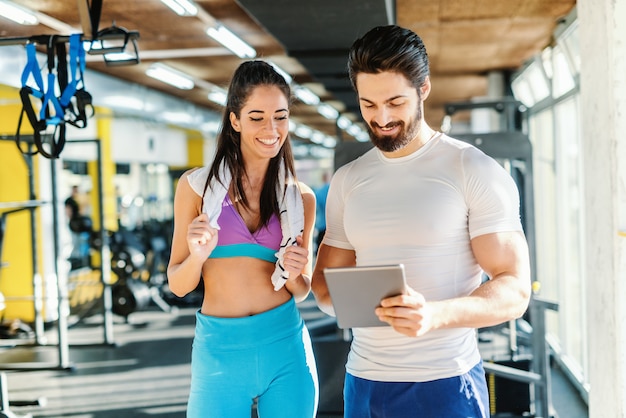 Bärtiger lächelnder persönlicher Trainer, der Frauenergebnisse des Trainings auf Tablette zeigt. Frau, die neben ihm steht und Tablette betrachtet. Innenraum des Fitnessraums.