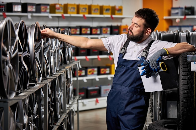 Bärtiger männlicher Automechaniker, der Felgen untersucht, das Sortiment in seinem Geschäft am Arbeitsplatz überprüft und Uniform trägt