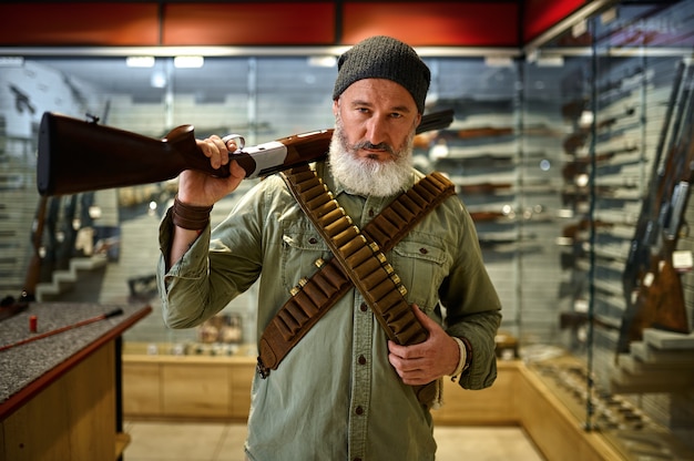 Foto bärtiger männlicher jäger mit gewehr und bandeliers im waffenladen. waffengeschäft, munitions- und munitionssortiment, waffenauswahl, schießhobby und lifestyle shooting