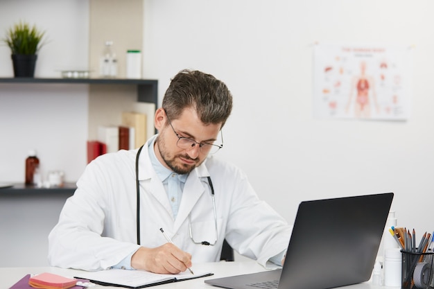 Bärtiger männlicher Therapeut im weißen medizinischen Kleid und in der Brille, die im medizinischen Schrank mit Stethoskop sitzen, Chirurg, der die Patientendokumente unter Verwendung des PCs analysiert