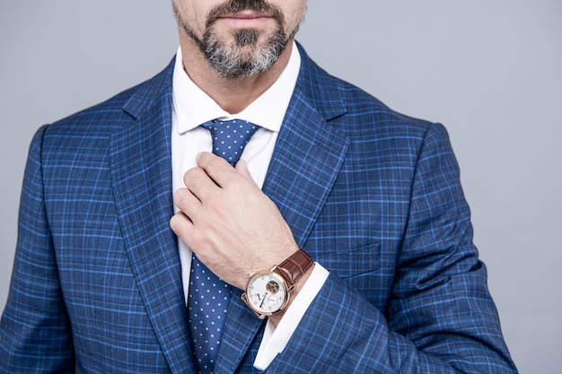 Bärtiger Mann beschnittene Ansicht fixieren Krawatte mit Business-Anzug und Luxusuhr grauer Hintergrund, Accessoire.
