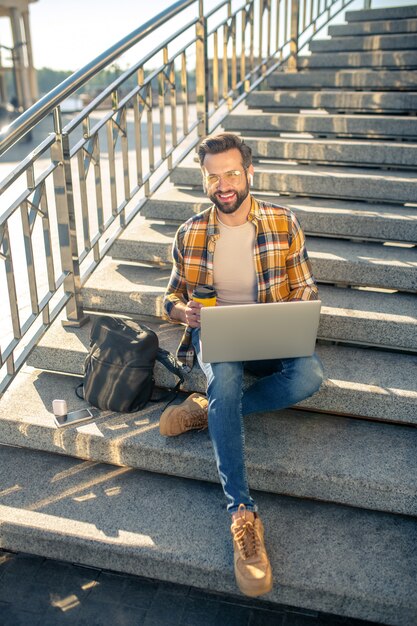 Bärtiger Mann, der an einem Laptop auf der Treppe arbeitet