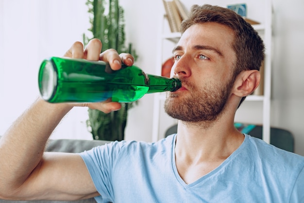 Bärtiger mann, der bier von der flasche trinkt, während zu hause auf sofa sitzen